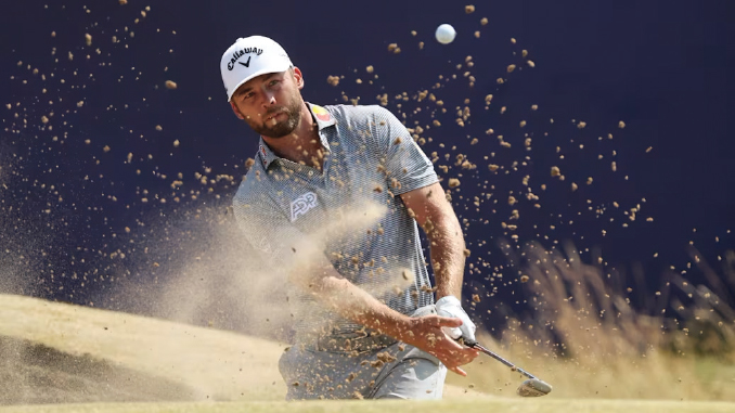 Male golfer in a white Callaway cap executes a bunker shot, sand spraying as he swings with his club.