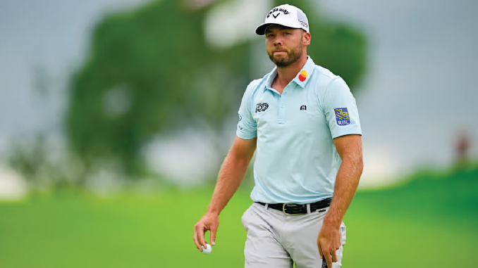Male golfer in a light blue polo and white cap walking on a lush golf course, holding a golf ball in his right hand.