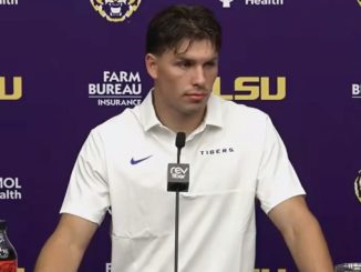 Male LSU Tigers football player at a podium in a press conference, wearing a white polo; purple backdrop with LSU logos and sponsor banners; Coca-Cola bottle on the table.
