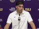Male LSU Tigers football player at a podium in a press conference, wearing a white polo; purple backdrop with LSU logos and sponsor banners; Coca-Cola bottle on the table.
