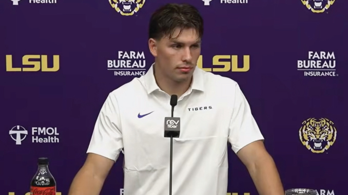 Male LSU Tigers football player at a podium in a press conference, wearing a white polo; purple backdrop with LSU logos and sponsor banners; Coca-Cola bottle on the table.