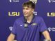 LSU football player in a purple polo at a press conference podium, backdrop with LSU logos and sponsor names, microphone in front, Coca‑Cola bottle on the table.