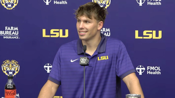 LSU football player in a purple polo at a press conference podium, backdrop with LSU logos and sponsor names, microphone in front, Coca‑Cola bottle on the table.