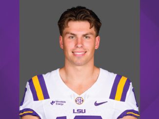 Portrait of a smiling LSU football player in a white jersey with purple and gold stripes, standing against a gray backdrop of the studio.