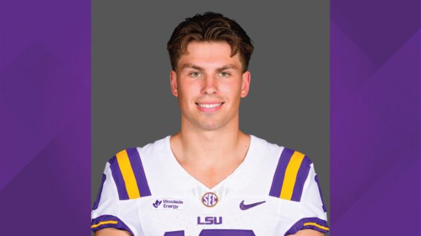 Portrait of a smiling LSU football player in a white jersey with purple and gold stripes, standing against a gray backdrop of the studio.