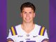 Portrait of a smiling LSU football player in a white jersey with purple and gold stripes, standing against a gray backdrop of the studio.