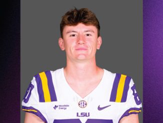 Portrait of a LSU football player in a white jersey with purple and gold stripes, standing against a gray backdrop