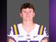 Portrait of a LSU football player in a white jersey with purple and gold stripes, standing against a gray backdrop
