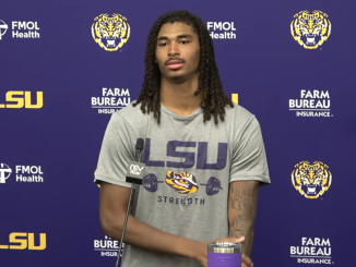 LSU football player with long dreadlocks at a press conference, wearing a gray 'LSU Strength' shirt, microphone at a podium against a purple backdrop with logos.