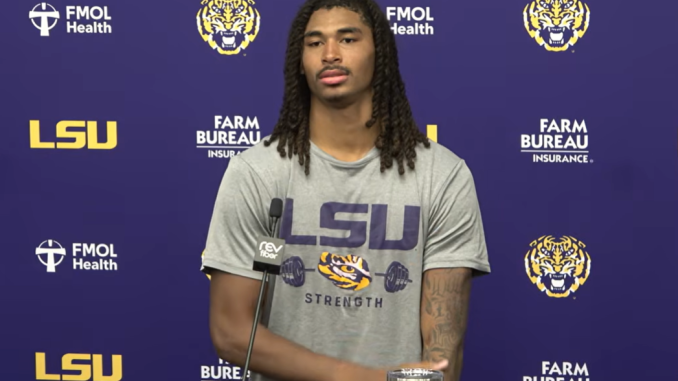LSU football player with long dreadlocks at a press conference, wearing a gray 'LSU Strength' shirt, microphone at a podium against a purple backdrop with logos.