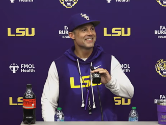 LSU coach in a purple hoodie at a press conference, speaking into a microphone with LSU logos on the backdrop behind him