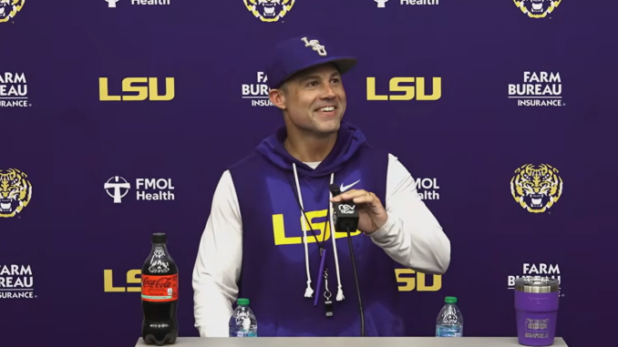 LSU coach in a purple hoodie at a press conference, speaking into a microphone with LSU logos on the backdrop behind him