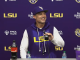 LSU coach in a purple hoodie at a press conference, speaking into a microphone with LSU logos on the backdrop behind him