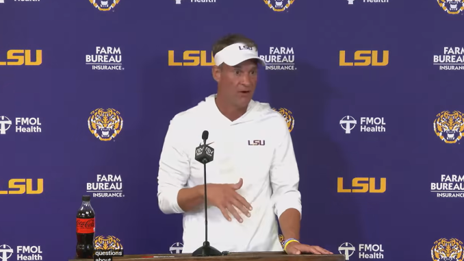 LSU football coach in a white hoodie and visor speaks at a podium during a press conference with a purple LSU backdrop and sponsor logos