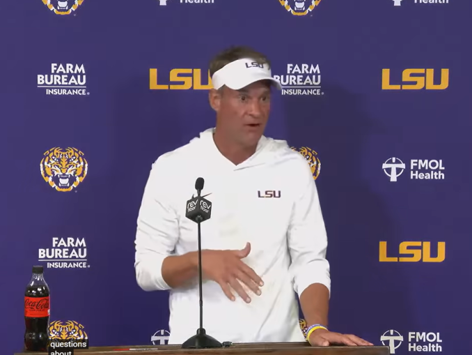 LSU football coach in a white hoodie and visor speaks at a podium during a press conference with a purple LSU backdrop and sponsor logos