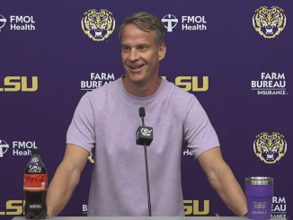 Man at a press conference behind a purple LSU backdrop, speaking into a microphone with a Coca‑Cola bottle on the podium's left and a purple tumbler on the right
