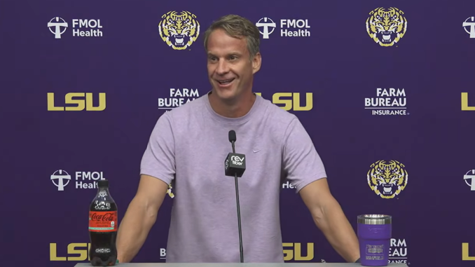 Man at a press conference behind a purple LSU backdrop, speaking into a microphone with a Coca‑Cola bottle on the podium's left and a purple tumbler on the right