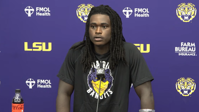 Athlete at a press conference in front of LSU and FMOL Health logos, wearing a Bandits shirt with a tiger logo; Coca-Cola bottle on the table.