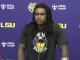 Athlete at a press conference in front of LSU and FMOL Health logos, wearing a Bandits shirt with a tiger logo; Coca-Cola bottle on the table.
