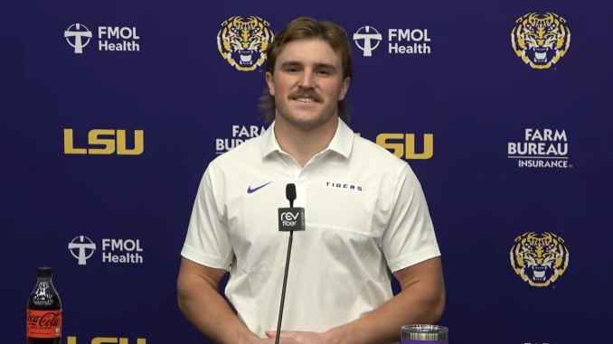 Male LSU athlete at a press conference, speaking into a mic while wearing a white polo against a branded backdrop with sponsors and logos.
