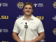 Male LSU athlete at a press conference, speaking into a mic while wearing a white polo against a branded backdrop with sponsors and logos.