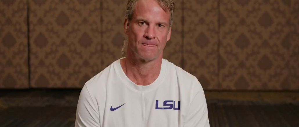 Man wearing a white LSU shirt sits facing the camera in a patterned room, likely during an interview.