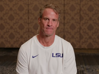 Man wearing a white LSU shirt sits facing the camera in a patterned room, likely during an interview.