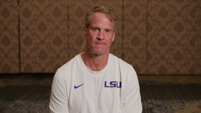 Man wearing a white LSU shirt sits facing the camera in a patterned room, likely during an interview.
