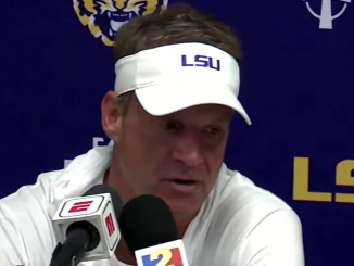 Man at a press conference wearing a white LSU visor, speaking into multiple microphones against a blue backdrop with LSU branding.