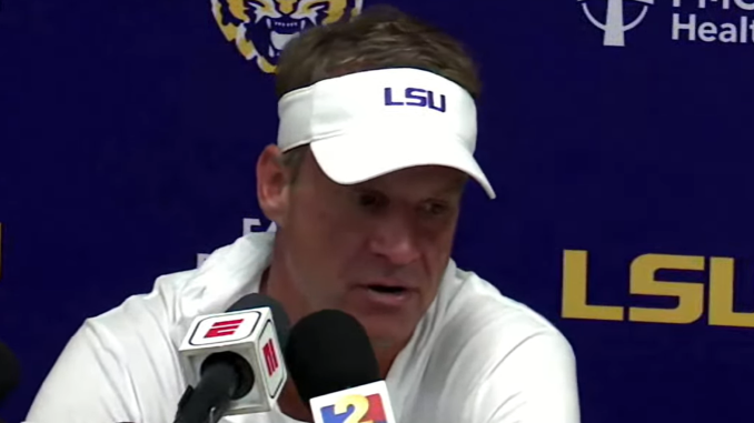 Man at a press conference wearing a white LSU visor, speaking into multiple microphones against a blue backdrop with LSU branding.