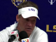 Man at a press conference wearing a white LSU visor, speaking into multiple microphones against a blue backdrop with LSU branding.