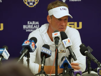 LSU football coach in a white visor and polo speaks at a press conference with multiple microphones in front of a purple LSU backdrop.