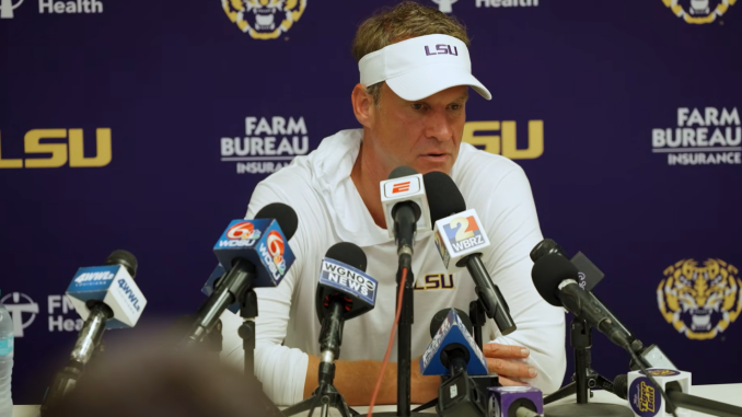 LSU football coach in a white visor and polo speaks at a press conference with multiple microphones in front of a purple LSU backdrop.