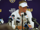 LSU football coach in a white visor and polo speaks at a press conference with multiple microphones in front of a purple LSU backdrop.