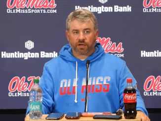 Ole Miss coach at a press conference, wearing a blue hoodie, speaking into a microphone with water and Coca-Cola bottles on the table.