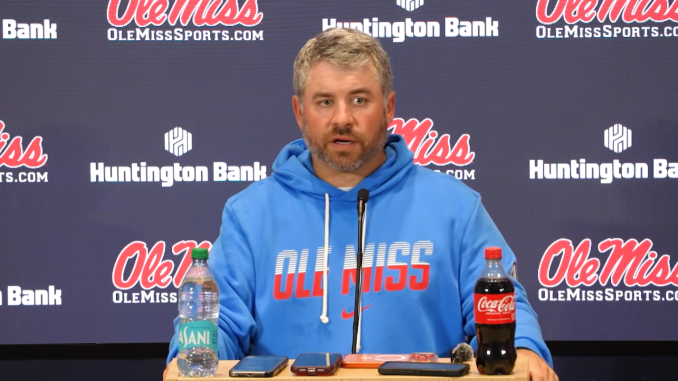 Ole Miss coach at a press conference, wearing a blue hoodie, speaking into a microphone with water and Coca-Cola bottles on the table.