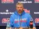 Ole Miss coach at a press conference, wearing a blue hoodie, speaking into a microphone with water and Coca-Cola bottles on the table.