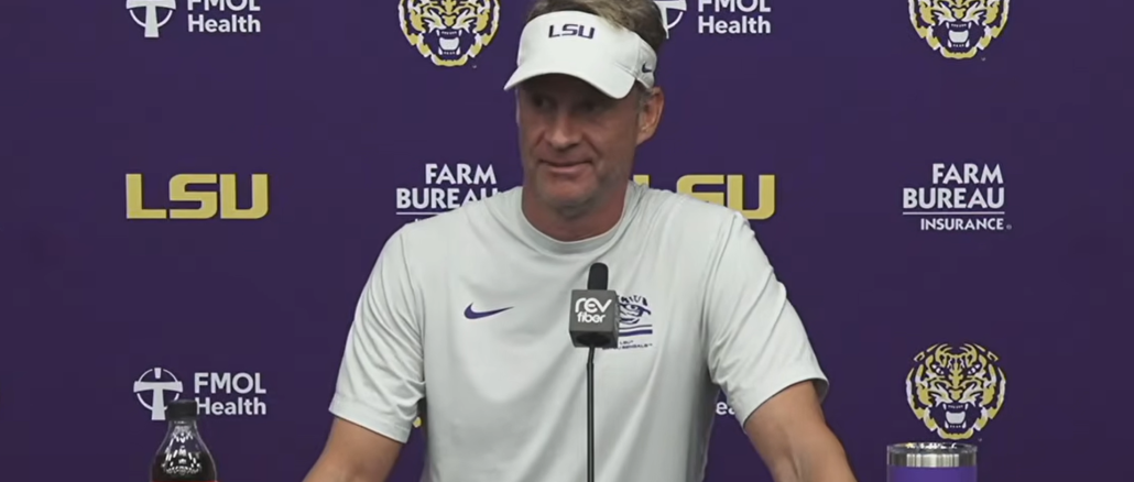 Man in a white LSU visor and shirt speaks at a press conference, purple backdrop with LSU and sponsor logos and a microphone in front.