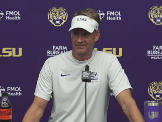 Man in a white LSU visor and shirt speaks at a press conference, purple backdrop with LSU and sponsor logos and a microphone in front.
