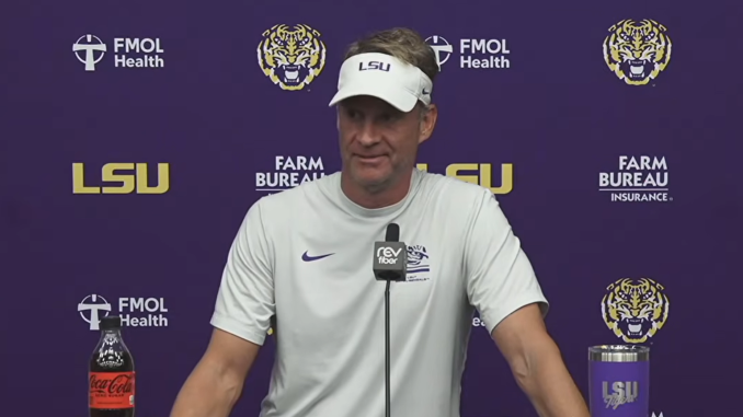 Man in a white LSU visor and shirt speaks at a press conference, purple backdrop with LSU and sponsor logos and a microphone in front.