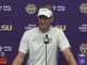 Man in a white LSU visor and shirt speaks at a press conference, purple backdrop with LSU and sponsor logos and a microphone in front.