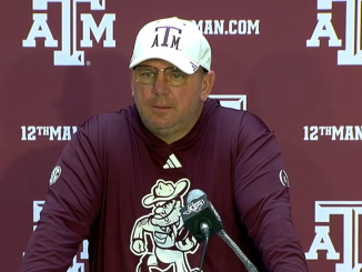 Man in maroon long-sleeve shirt and white A&M cap speaks at a microphone during a Texas A&M press conference backdrop.