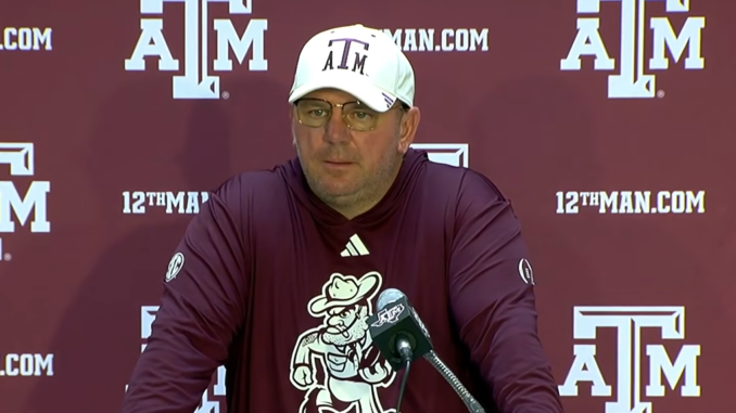 Man in maroon long-sleeve shirt and white A&M cap speaks at a microphone during a Texas A&M press conference backdrop.