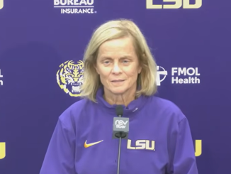Woman in a purple LSU sweatshirt speaks at a microphone during a press conference with LSU logos in the background.