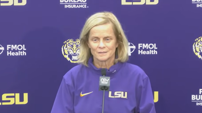 Woman in a purple LSU sweatshirt speaks at a microphone during a press conference with LSU logos in the background.