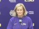 Woman in a purple LSU sweatshirt speaks at a microphone during a press conference with LSU logos in the background.