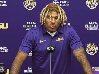 LSU football player at a press conference, wearing a purple polo with LSU logo, speaking into a microphone against a purple backdrop with sponsor logos.