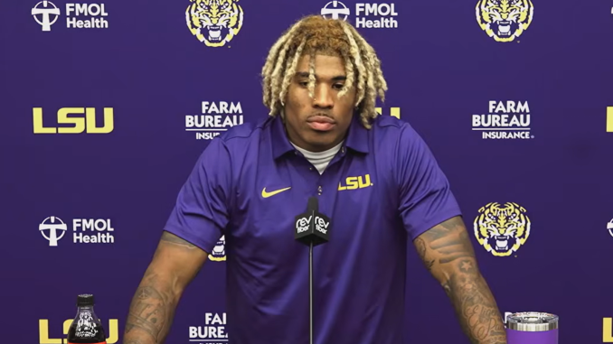 LSU football player at a press conference, wearing a purple polo with LSU logo, speaking into a microphone against a purple backdrop with sponsor logos.