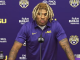 LSU football player at a press conference, wearing a purple polo with LSU logo, speaking into a microphone against a purple backdrop with sponsor logos.
