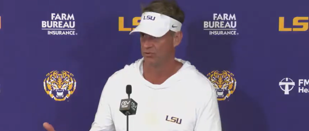 LSU coach at a press conference podium, wearing a white hoodie and visor, speaking against a purple LSU backdrop with logos.